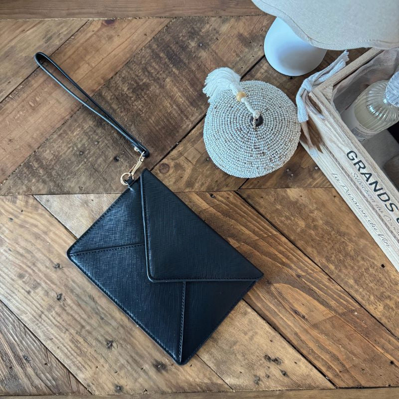 Pochette enveloppe en simili cuir noir avec dragonne, posée à plat sur un parquet en chevrons.