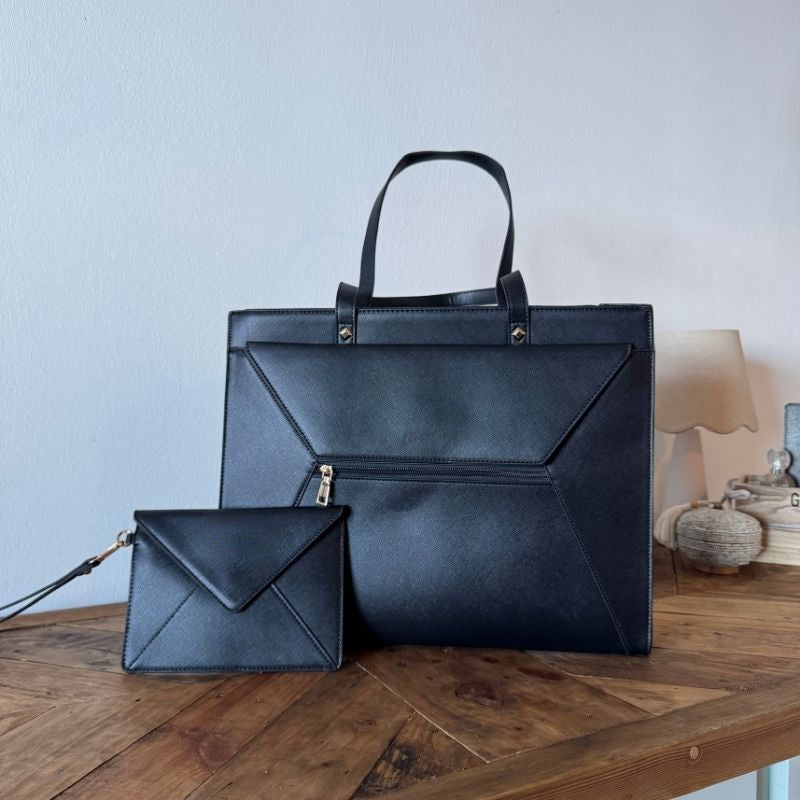 Sac à main pour ordinateur noir vu de face sur une console en bois, une pochette enveloppe noire à dragonne posée devant.