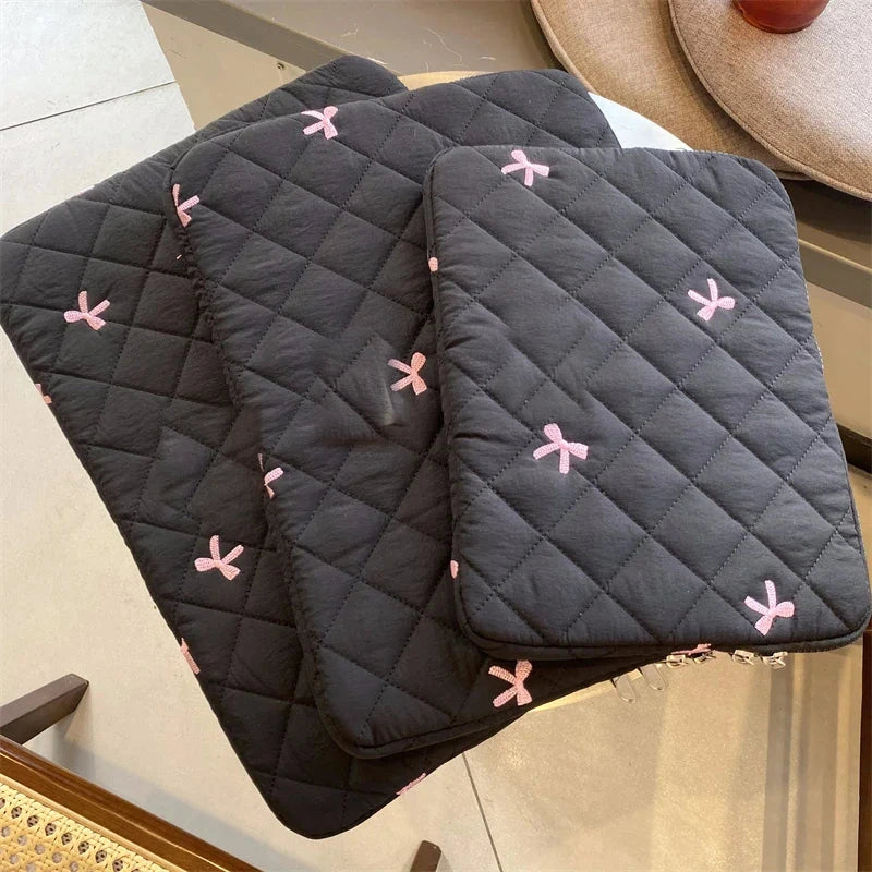 Trois housses matelassées noires pour ordinateur portable, brodées de petits nœuds roses, posées en éventail sur une table.