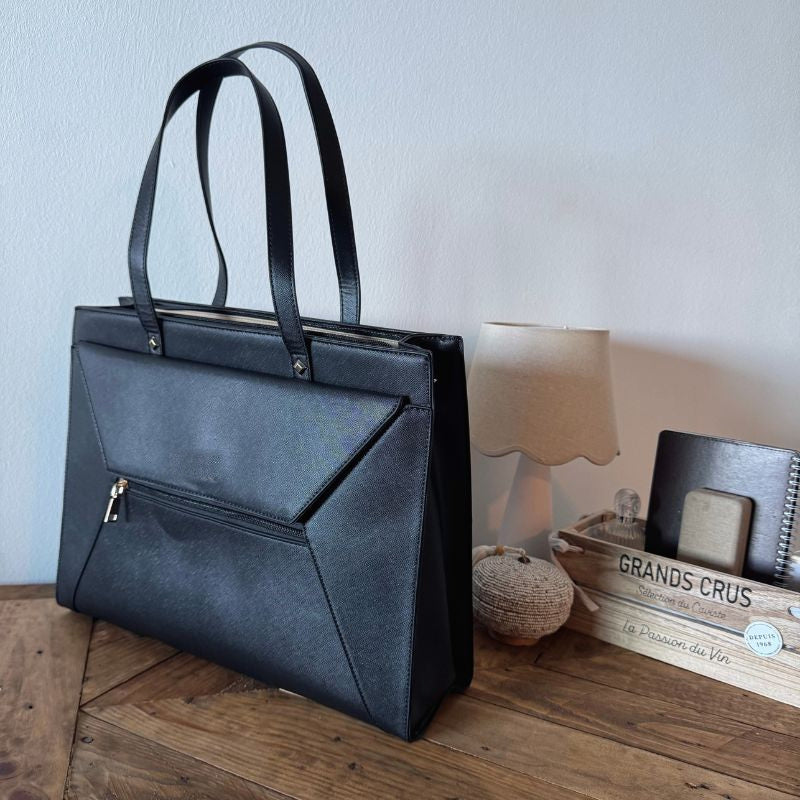 Sac à main pour ordinateur en simili cuir noir vu de trois quarts sur un parquet, rabat enveloppe et poche avant zippée.